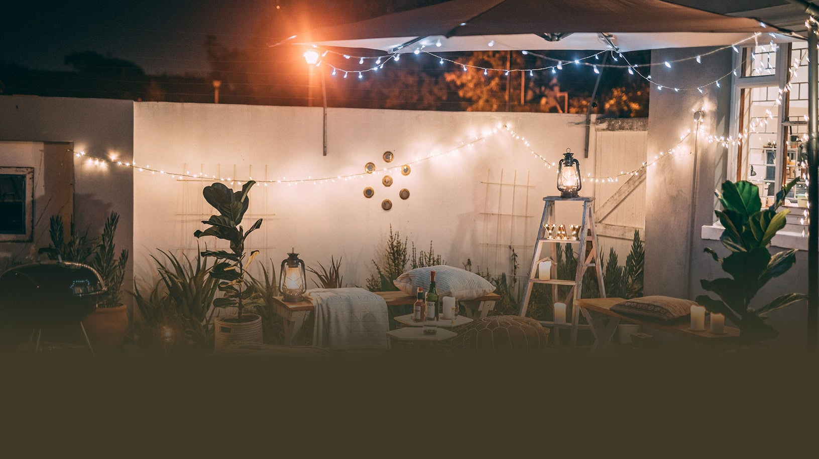 Cozy outdoor patio at night, adorned with string lights and lanterns. Cushions and plants create an inviting, warm ambiance.