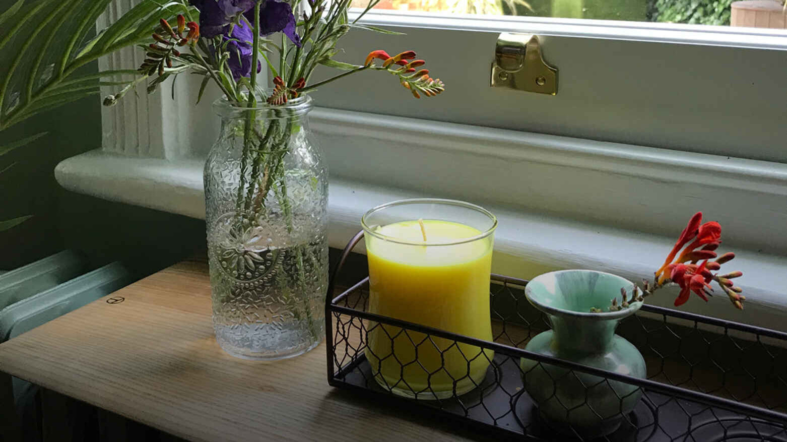 A glass vase with purple and red flowers, a yellow candle, and a small ceramic vase sit on a wooden windowsill, creating a cozy, serene atmosphere.
