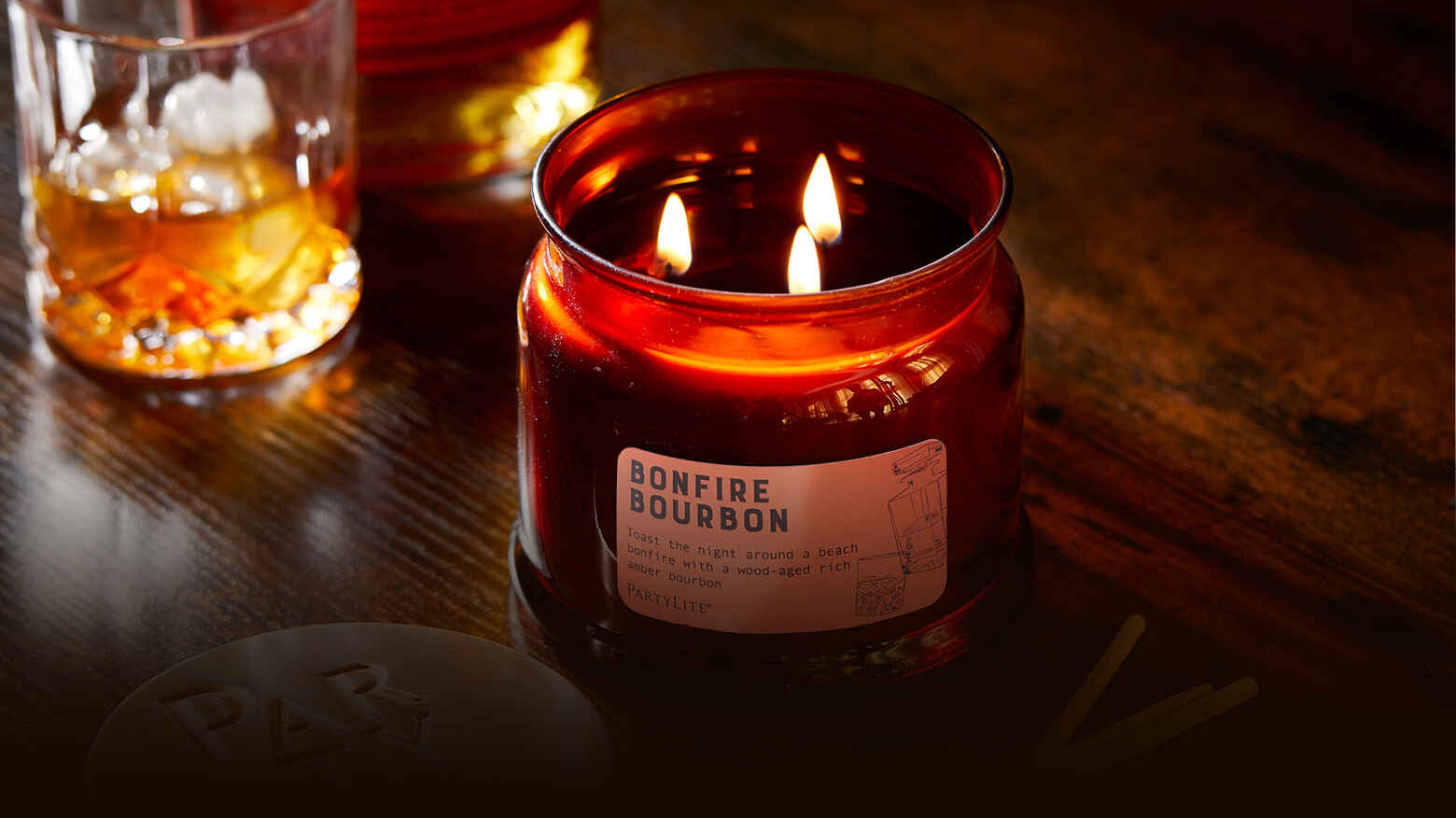 A warm amber candle labeled "Bonfire Bourbon" burns beside a glass of whiskey on a wooden table, creating a cozy, intimate atmosphere.