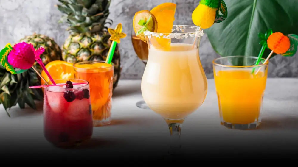 A variety of colorful cocktails on a table, each garnished with fruit and umbrellas. Background includes pineapples and a large green leaf, conveying a tropical vibe.