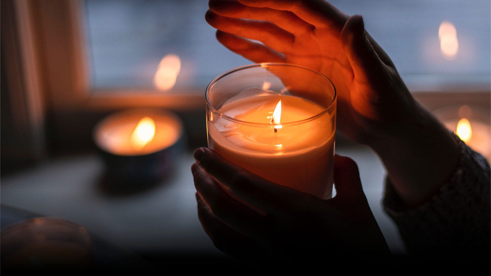 A lit jar candle against a person hands on a table