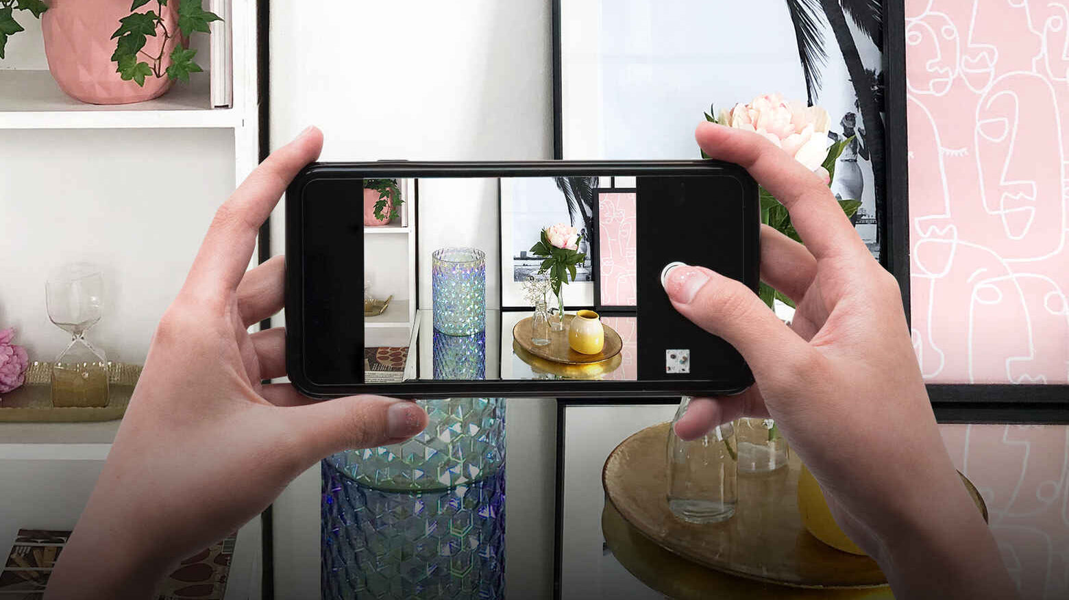 Woman using her phone to take a picture of a place setting consisting of a Shimmering Facets Hurricane candle holder and a vase with a carnation.
