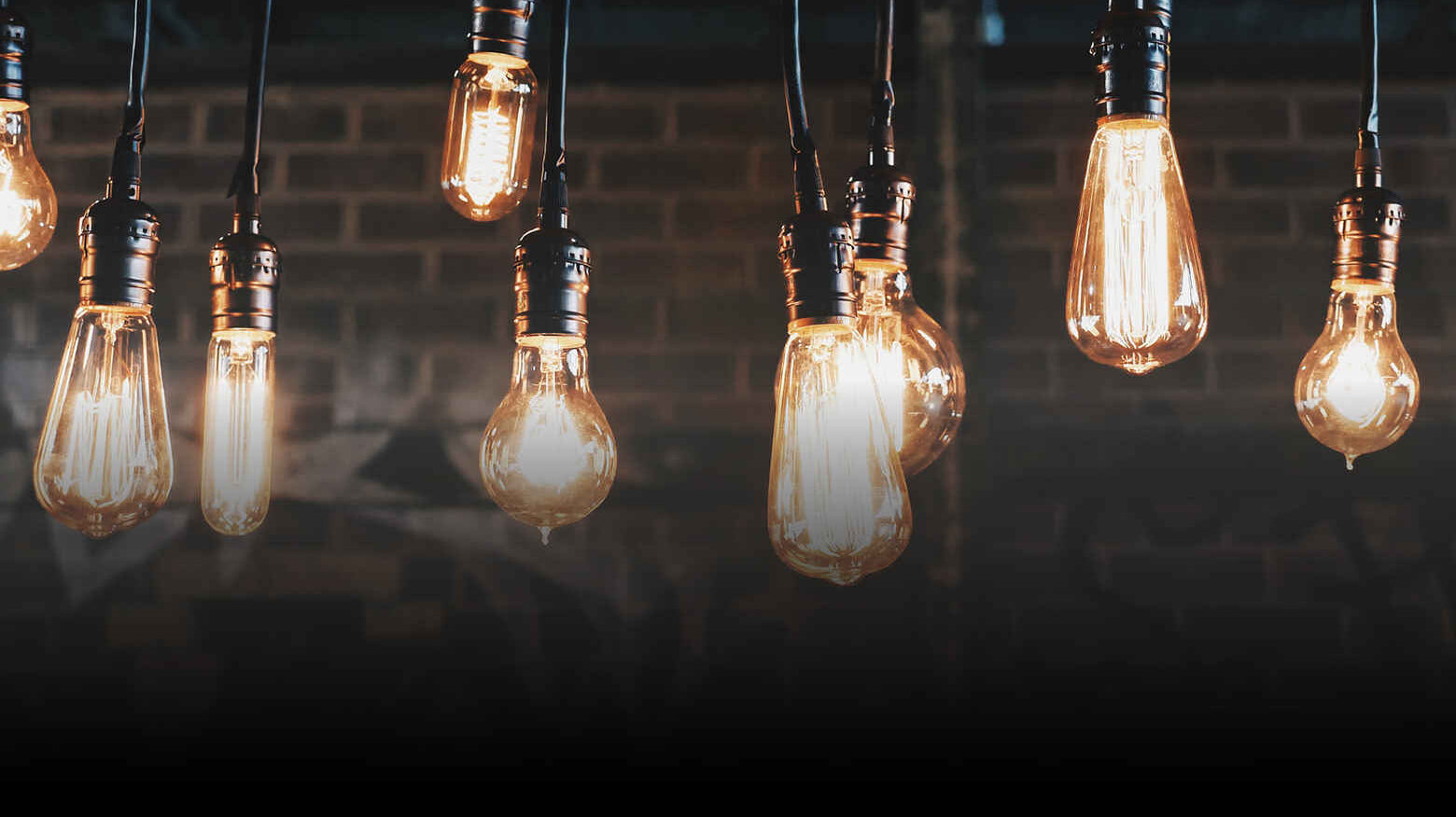 Hanging vintage Edison bulbs cast a warm glow against a dark brick wall, creating a cozy, nostalgic ambiance. The image conveys warmth and rustic charm.