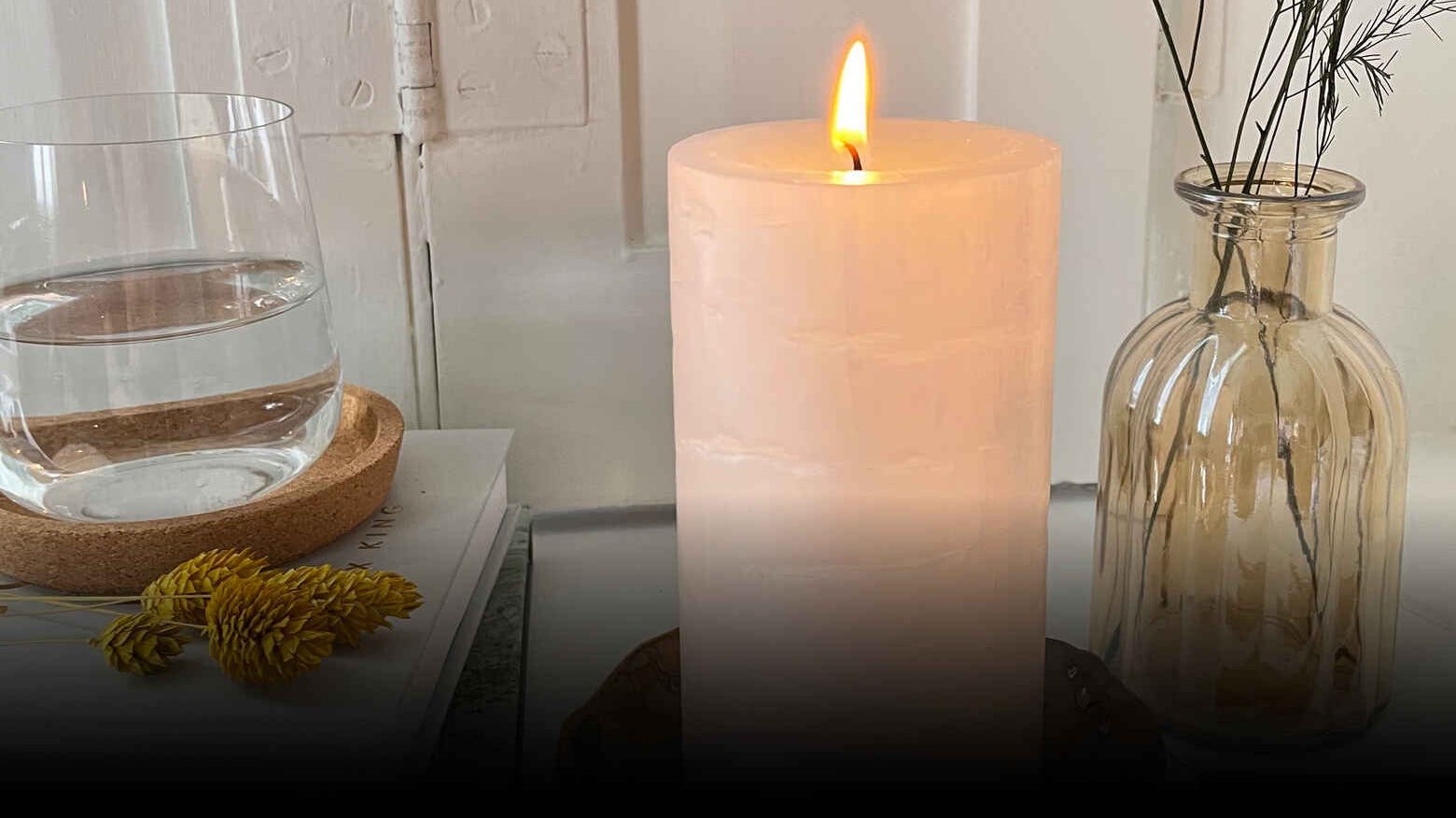 Pillar candle shown lit against a clear vase and a glass of water.