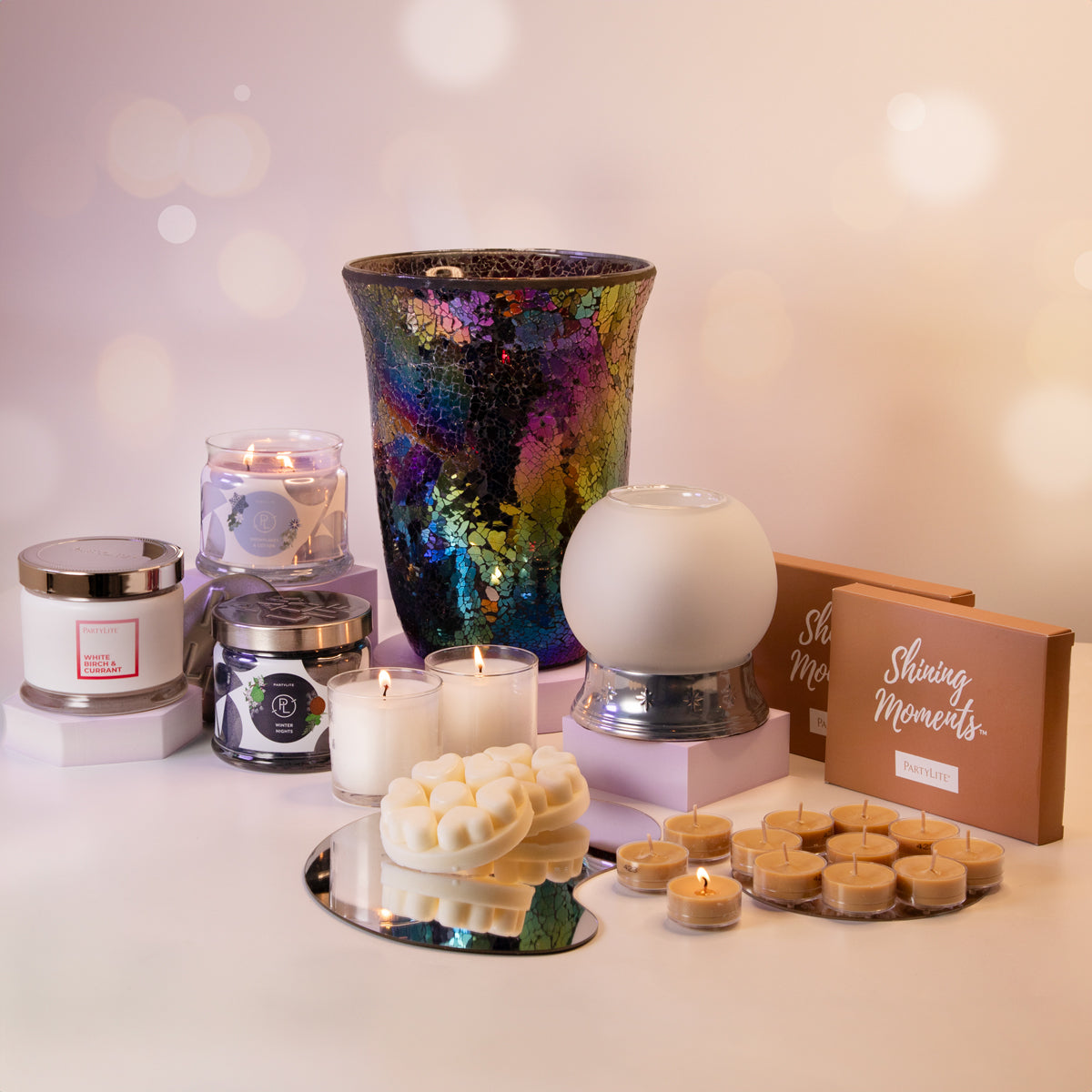 A collection of PartyLite gift items including various candles in jars and holders, tealights, a scent glow warmer, and wax melt cups, displayed on a table with a decorative background.