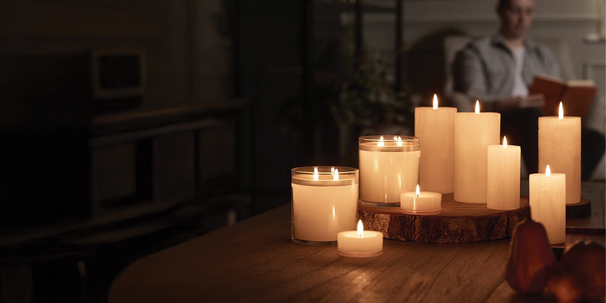 Multiple Glolite jar, pillar and tealight candles in a dimly lit space with man reading book in a blurred background
