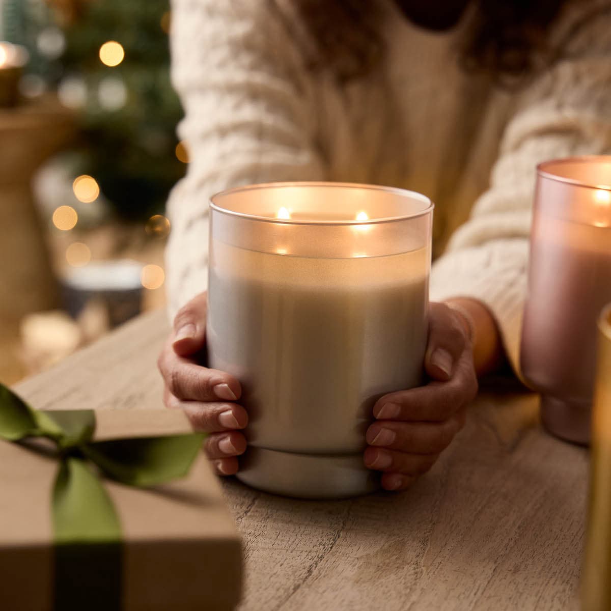 Iced Snowberries 2-Wick Specialty Jar Candle in a gold frosted glass lit, being held in a lady hands on a wooden surface.