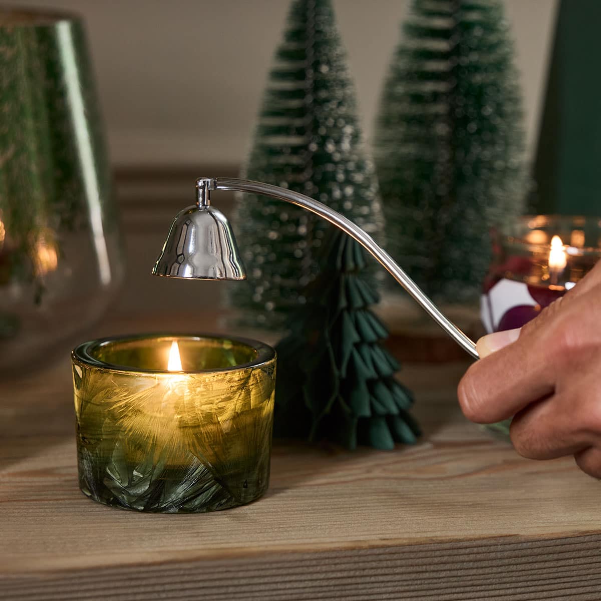 Forest Mist Large Tealight Holder lit, with a snuffer being held just above the holder with decorative Christmas trees in the background.
