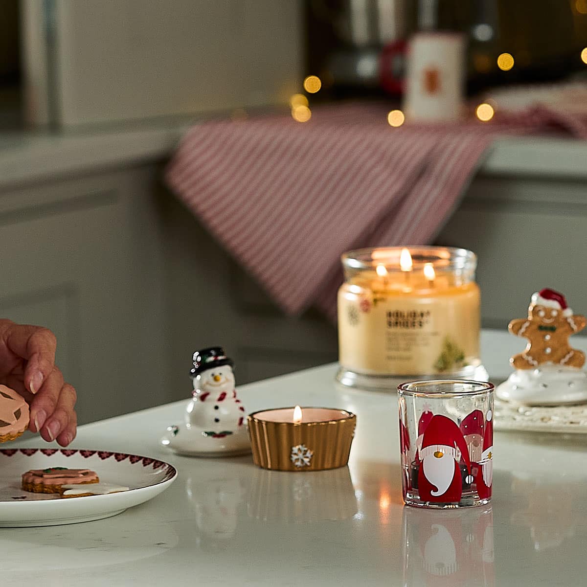 Gnome-for-the-Holidays Tealight/Votive Holder shown with Snowman & Gingerbread tealight holders with a lit 3-wick jar and a plate of cookies on a kitchen counter.