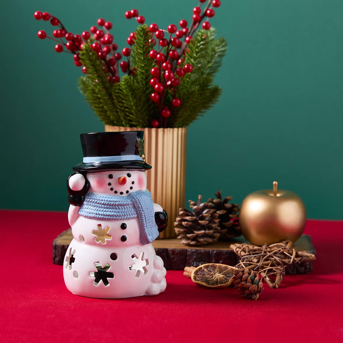 Decorative snowman with a vase of red berries and Christmas decorations on a red surface.