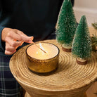 Whiskey & Oak 3-Wick Ripple Jar Candle being lit with a long match stick by a lady hand on a wood surface with decorative glittered tips Christmas Trees.