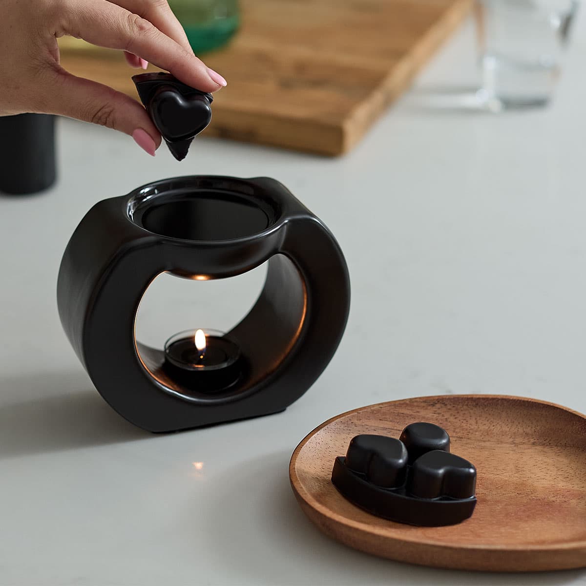Black Gaia Fragrance Warmer shown with a lit tealight, with black heart shaped wax melts on a wooden tray.