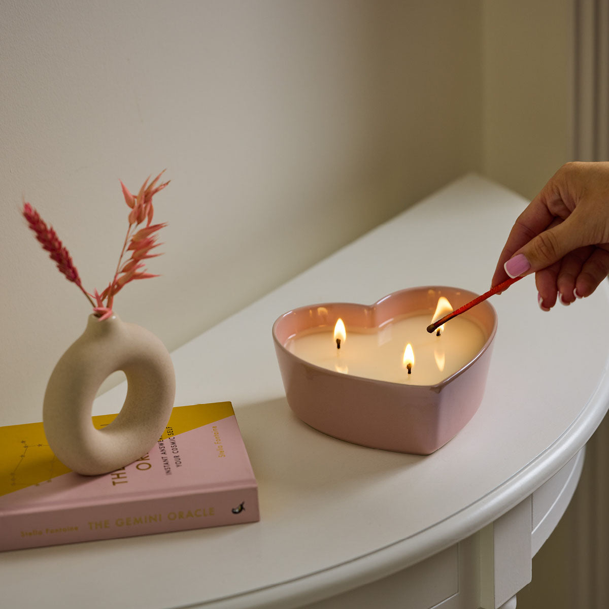 Iced Snowberries Heart shaped ceramic pink jar candle showing the wicks being lit with a cream vase on a book behind it on a white surface.