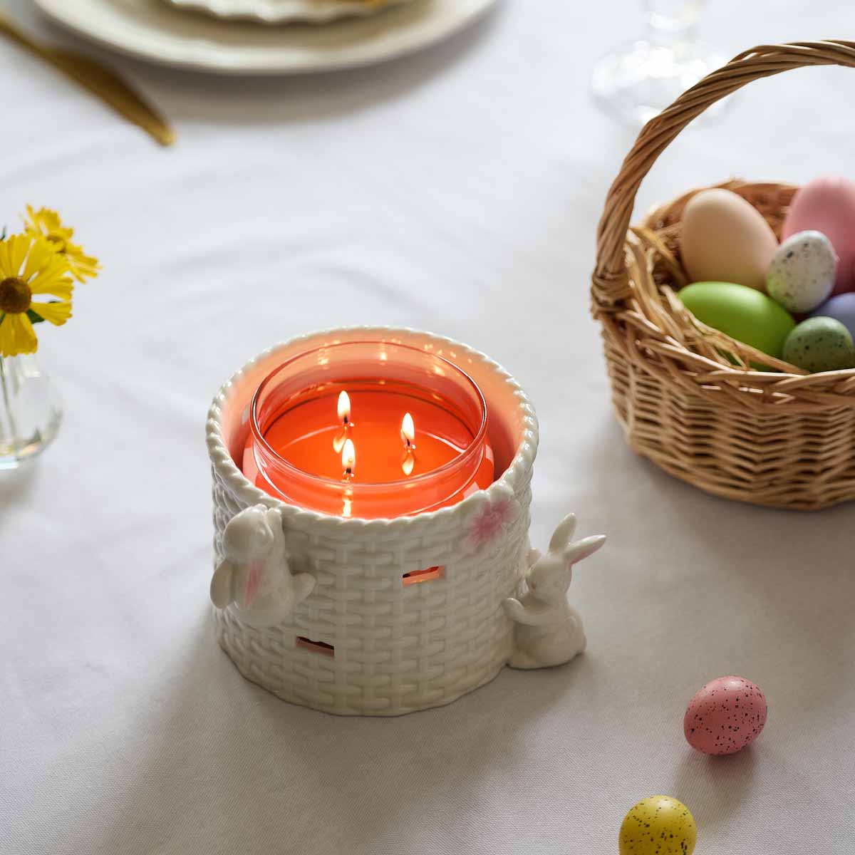 Easter Basket Candle Holder with Bunny Rabbits shown with a lit 3-wick jar candle next to a basket of Easter Eggs.