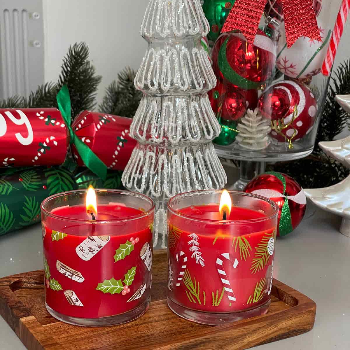 A pair of holiday-themed mini wax in clear jars with Christmas decorations, lit on a wooden table with Christmas items in the background.