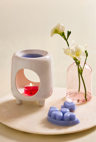 Mini Chiminea Fragrance Warmer with melt and burning tealight next to a melt and glass bud vase with flowers on a round stand