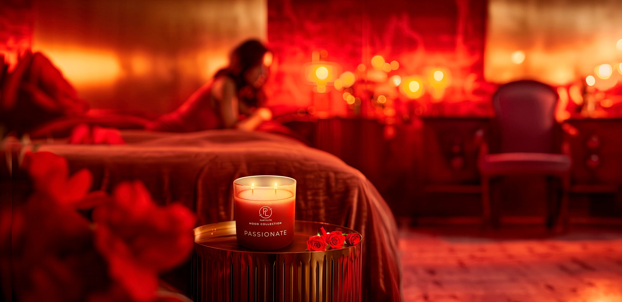 Passionate Mood Jar Candle on a round table with roses next to the jar with a blurred background in red with bed and lady on it in the background