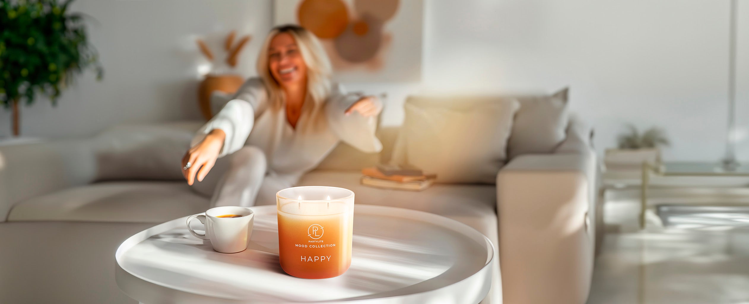 Happy Mood Jar Candle on a light colored table with a hot drink next to it and a lady sitting on a light colored sofa in the background