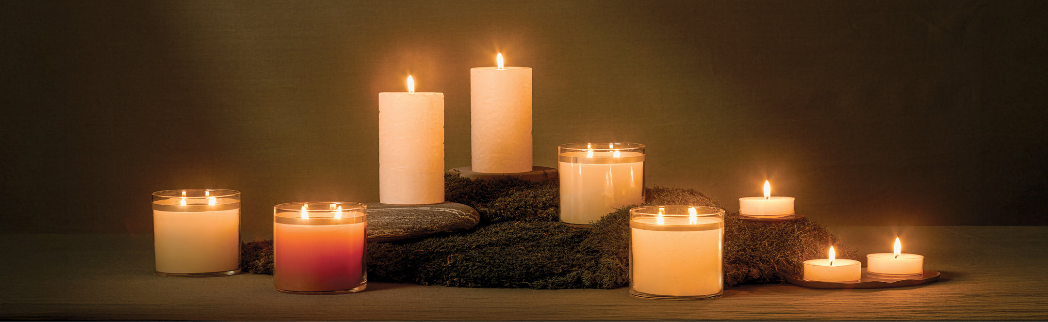 Row of lit GloLite jar, pillar and tealight candles on a dark background with some set upon stones.