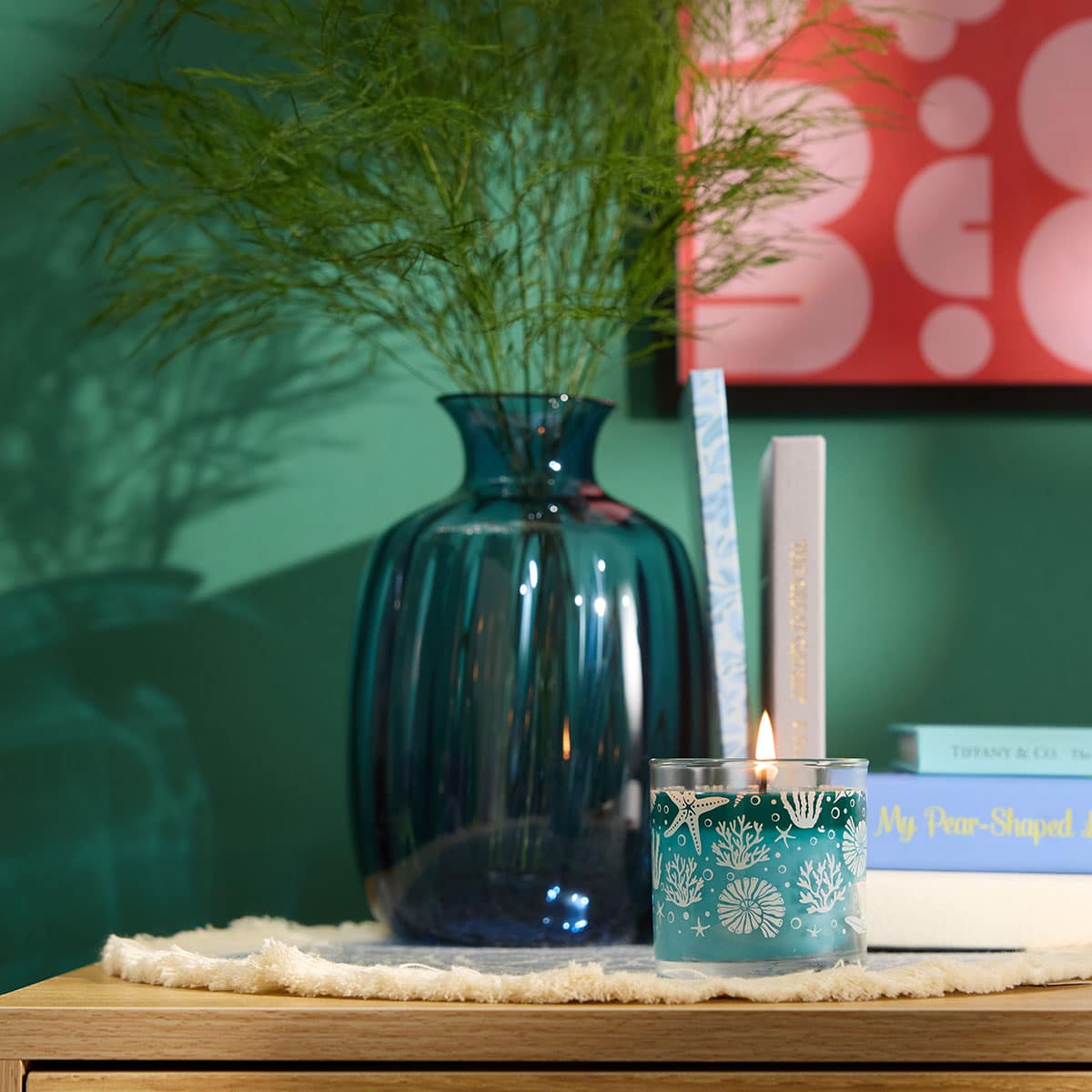 Salty Seascape Mini Jar Candle shown with a clear blue vase with flowers on a cream table mat on a wooden surface with books in the background against a green wall