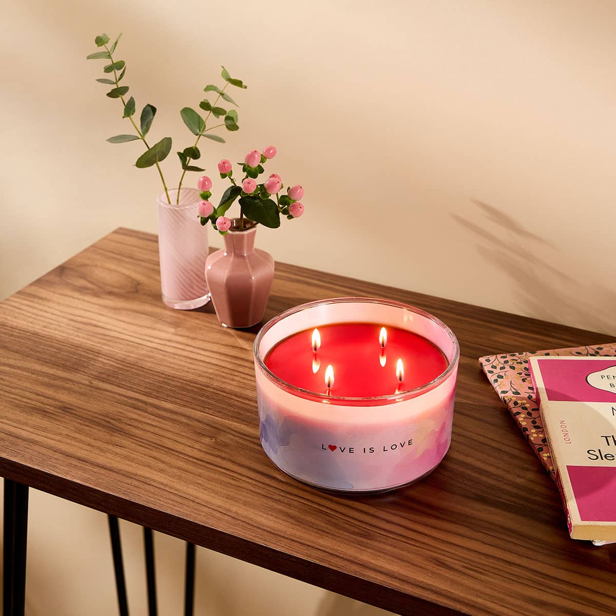 Love is Love 4-wick Jar Candle shown on a wooden table top with a vase of flowers and books next to it, against a beige background