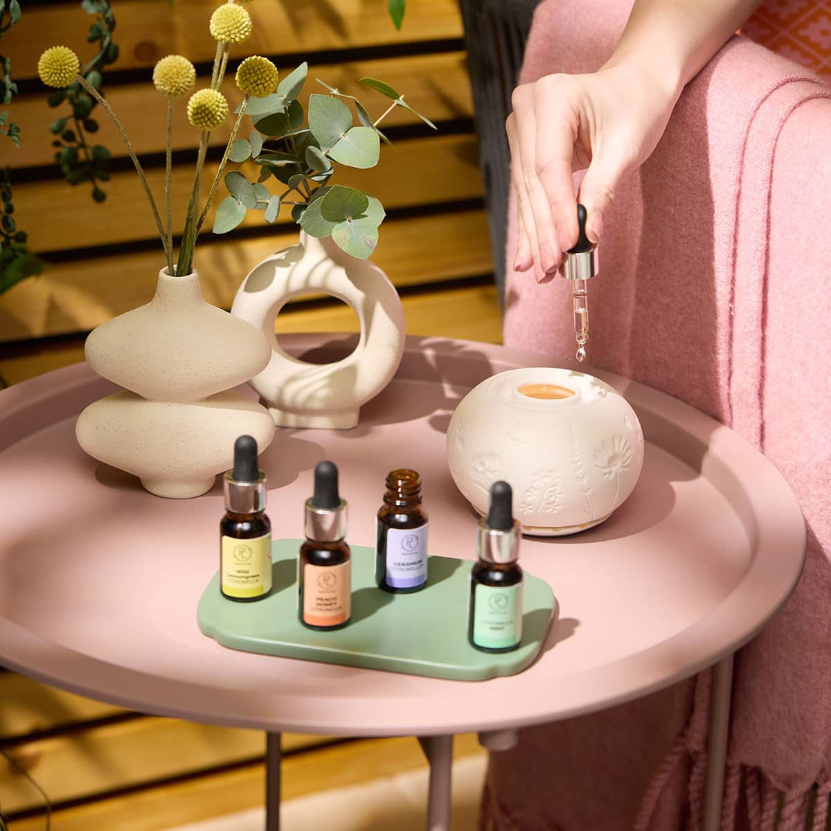 Four different Citronella Fragrance Oils on a Green tray shown with a Fragrance Warmer on a pink table and a hand adding oil into the top of the fragrance warmer with decorative vase of flowers next to in