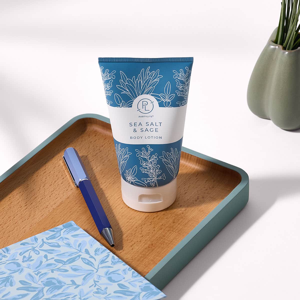Sea Salt and Sage body lotion shown on a wooden tray next to a pen and notepad on a white background