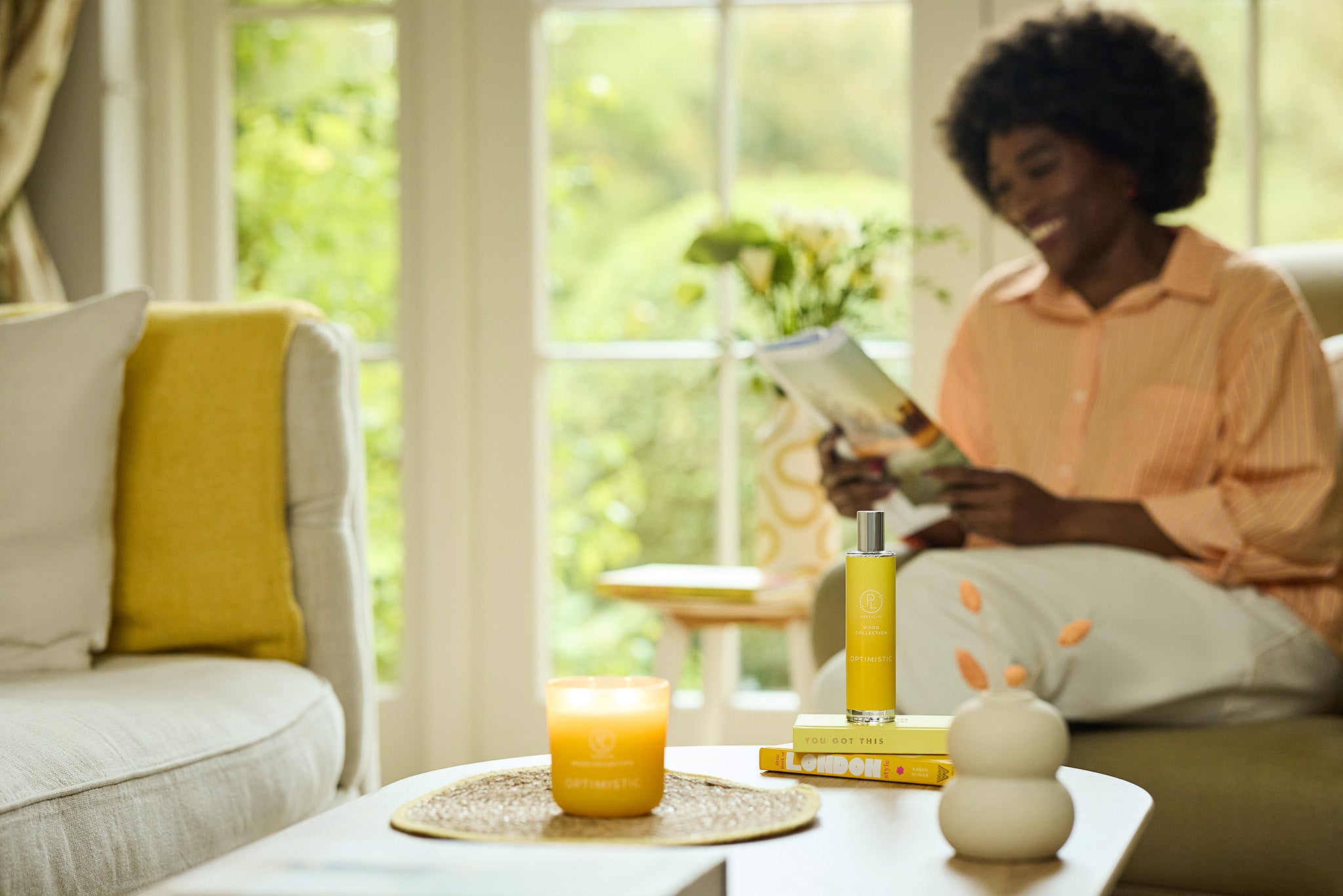 Optimistic Mood Jar and Room spray shown on a table with a lady sitting in the background smiling with a magazine in her hands