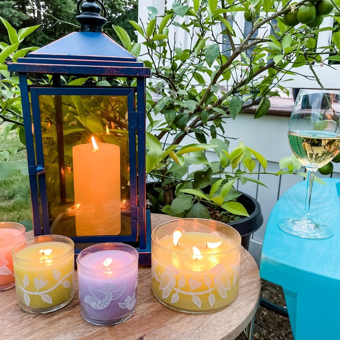 Various GlotLite Citronella jar candles, in Peach, Lilac and yellow colors, with a GloLite pillar candle ina blue lantern on a round wooden table with plants in the background.
