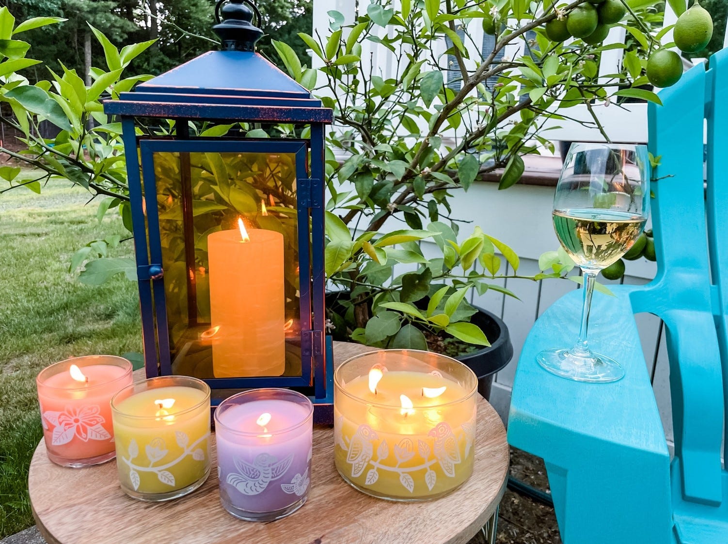 Various GlotLite Citronella jar candles, in Peach, Lilac and yellow colors, with a GloLite pillar candle ina blue lantern on a round wooden table with plants in the background.