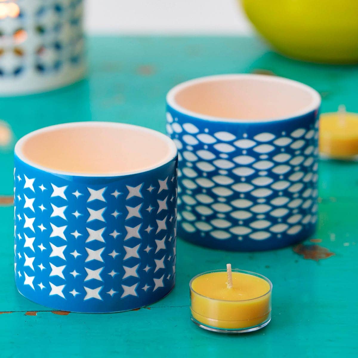 Grecian Tealight Candle Holder Pair on a green surface with a single yellow tealight in front of them.