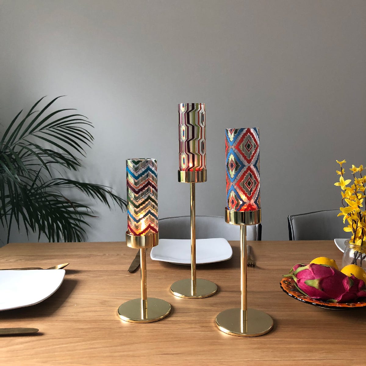 Tribal Chic Raised Tealight Holder Trio colorful pattern cup on a gold stand on a wooden dining table with plate settings.