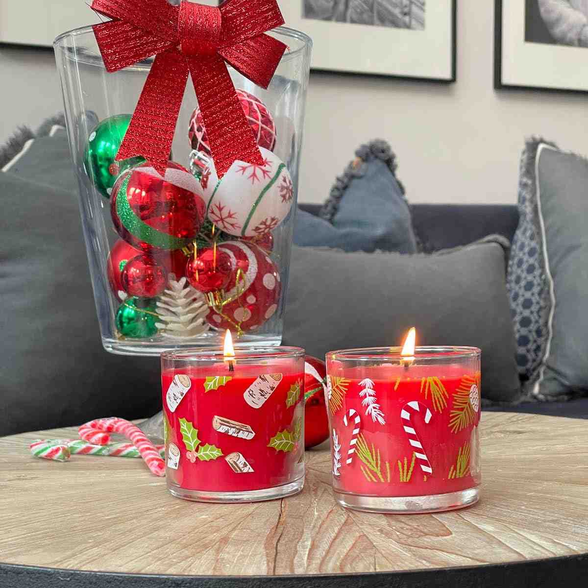 A pair of holiday-themed mini wax in clear jars with Christmas decorations lit on a wooden table with a jar of Christmas baubles