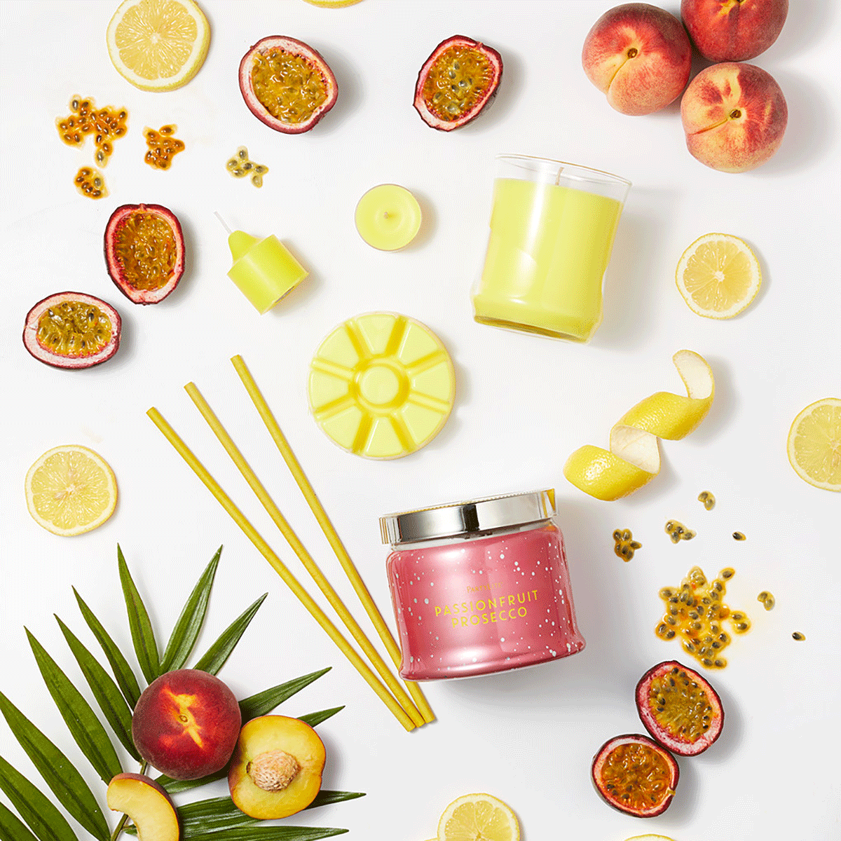 Passionfruit Prosecco 3-wick jar candle, fragrance sticks, escential jar, votive, tealight and wax melt, surrounded by slices of lemon and peaches against a white background.