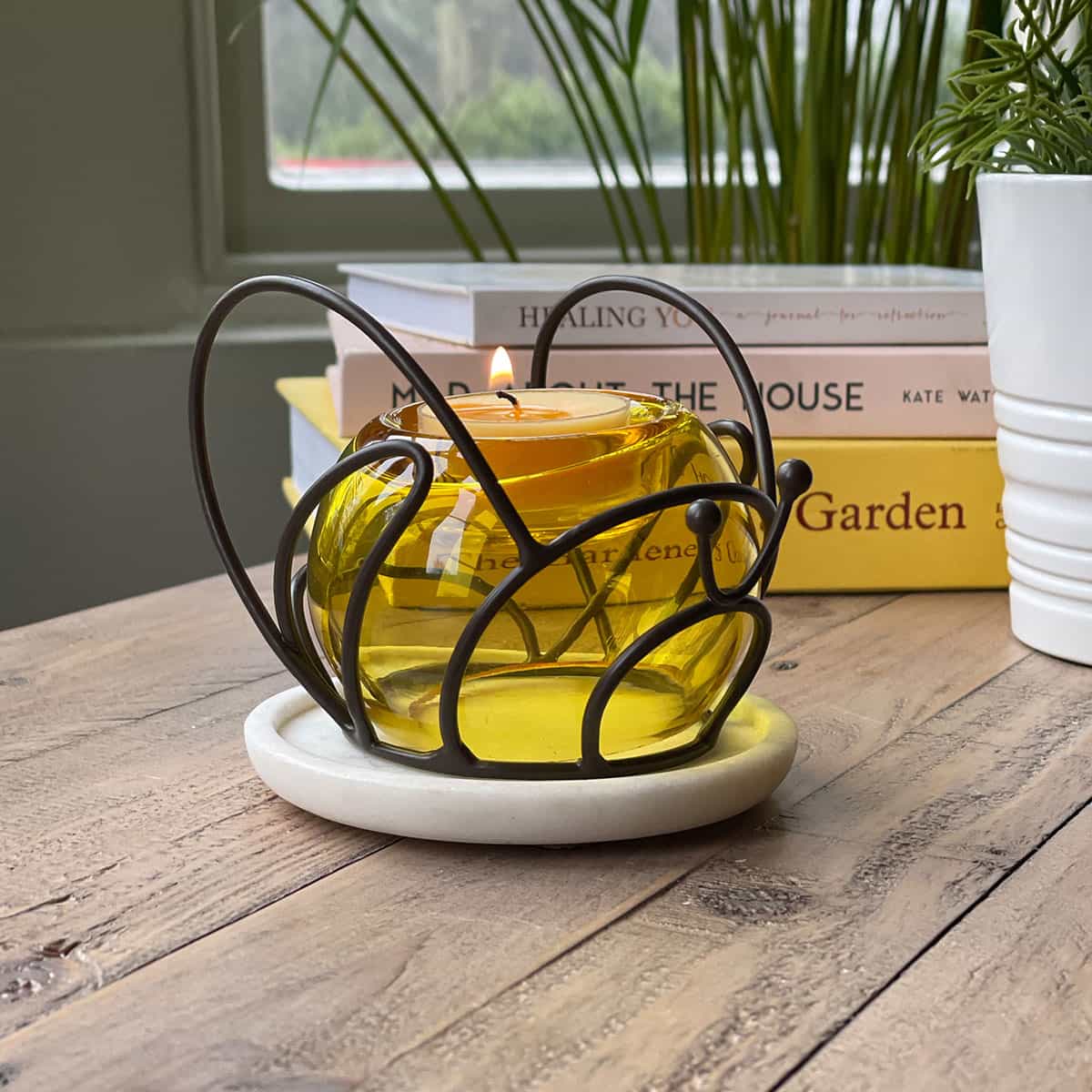 A dark metal tealight holder designed to resemble a bee, with a yellow glass globe for the body on a wooden table with books in the background.