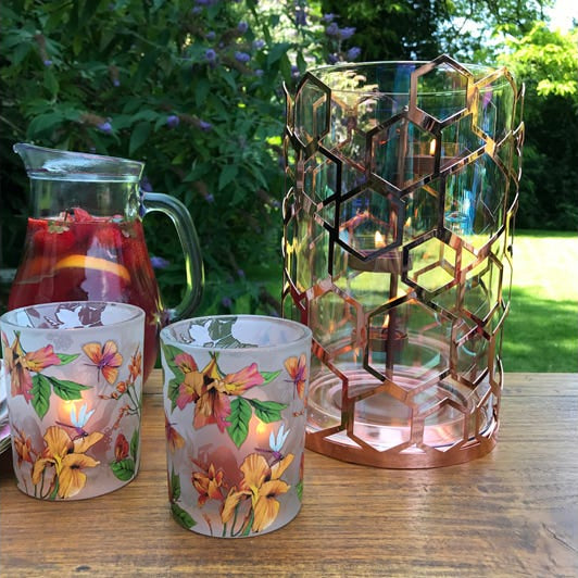 Afternoon Breeze Tealight Candle Holder Pair shown with lit tealights and a jug of juice, and rose gold hurricane on a wooden table with grass and trees in the background.