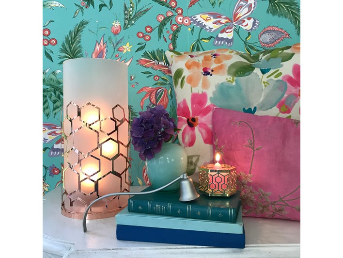 Frosted Honeycomb Hurricane with rose gold colored metal hexagon shapes with a white frosted insert, with lit candles on a shelf with colorful wallpaper and books.