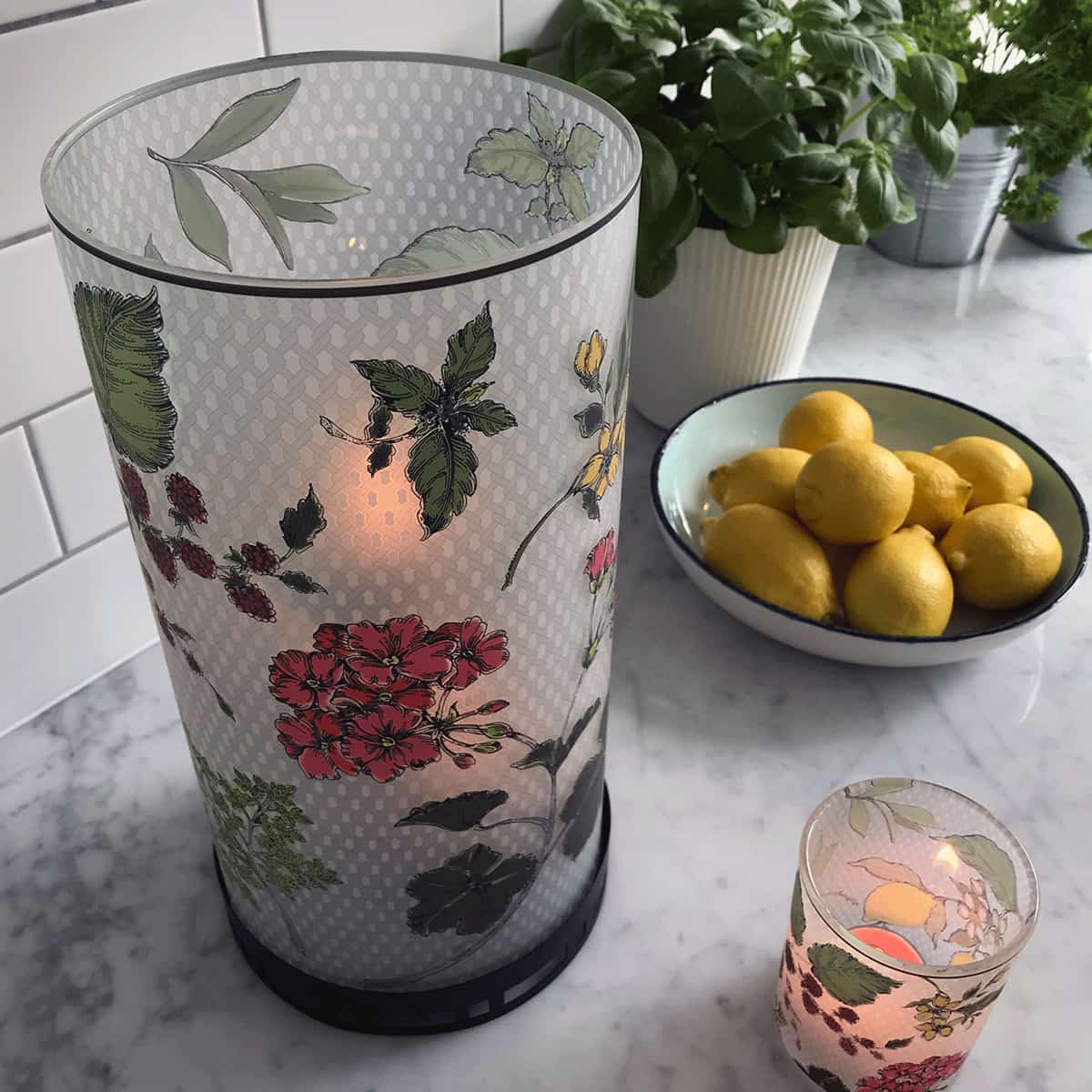 Fresh Garden Mix Hurricane with red and yellow colored flowers and green leaves, next a lit tealight holder and a bowl of lemons.