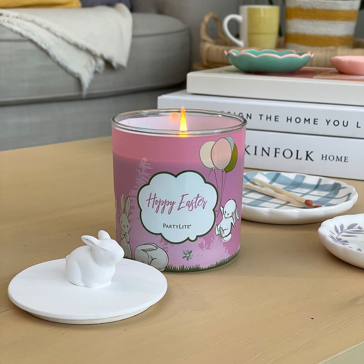 A jar candle with a pink label featuring Easter-themed graphics, including a bunny and balloons, and a white lid with a rabbit knob, on a wooden surface with books and plates in the background.