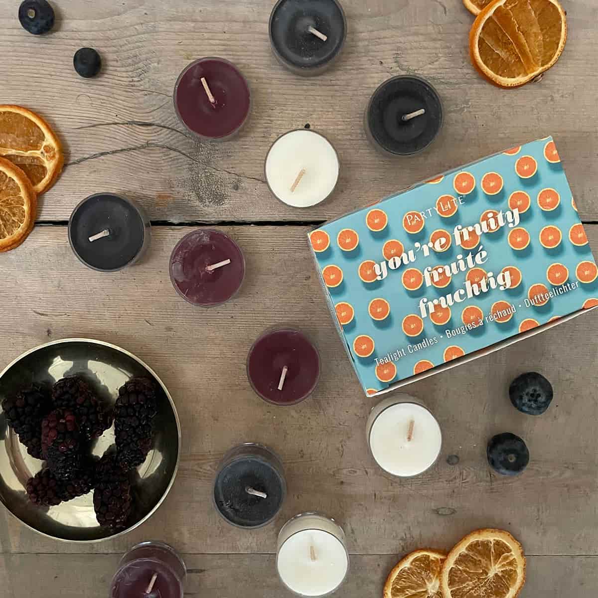 You're Fruity 12-Piece Tealight Sampler different colored tealights against a wooden surface with slices of oranges.