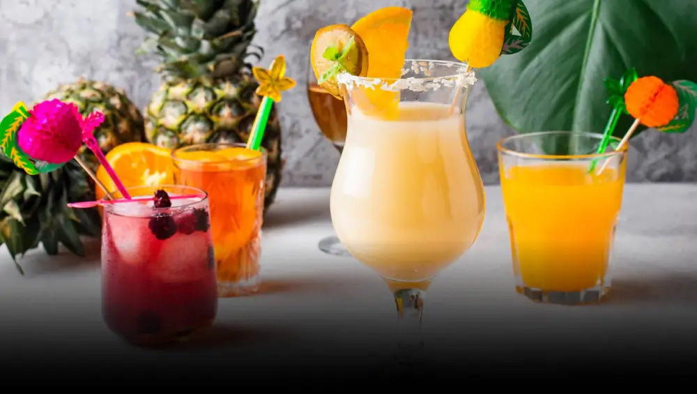 A variety of colorful cocktails on a table, each garnished with fruit and umbrellas. Background includes pineapples and a large green leaf, conveying a tropical vibe.