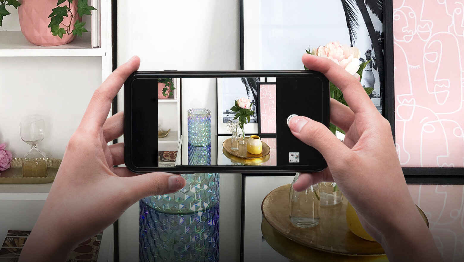 Woman using her phone to take a picture of a place setting consisting of a Shimmering Facets Hurricane candle holder and a vase with a carnation.