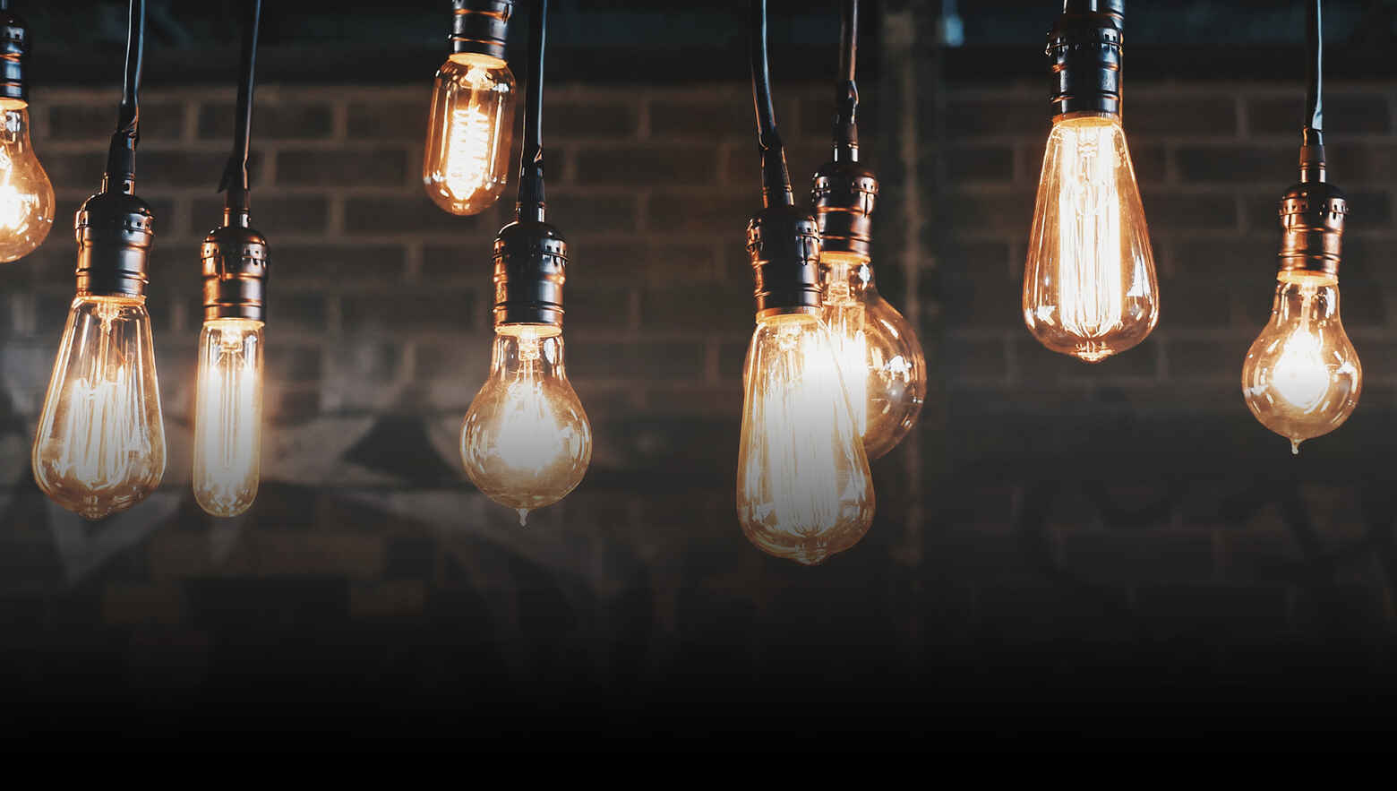 Hanging vintage Edison bulbs cast a warm glow against a dark brick wall, creating a cozy, nostalgic ambiance. The image conveys warmth and rustic charm.