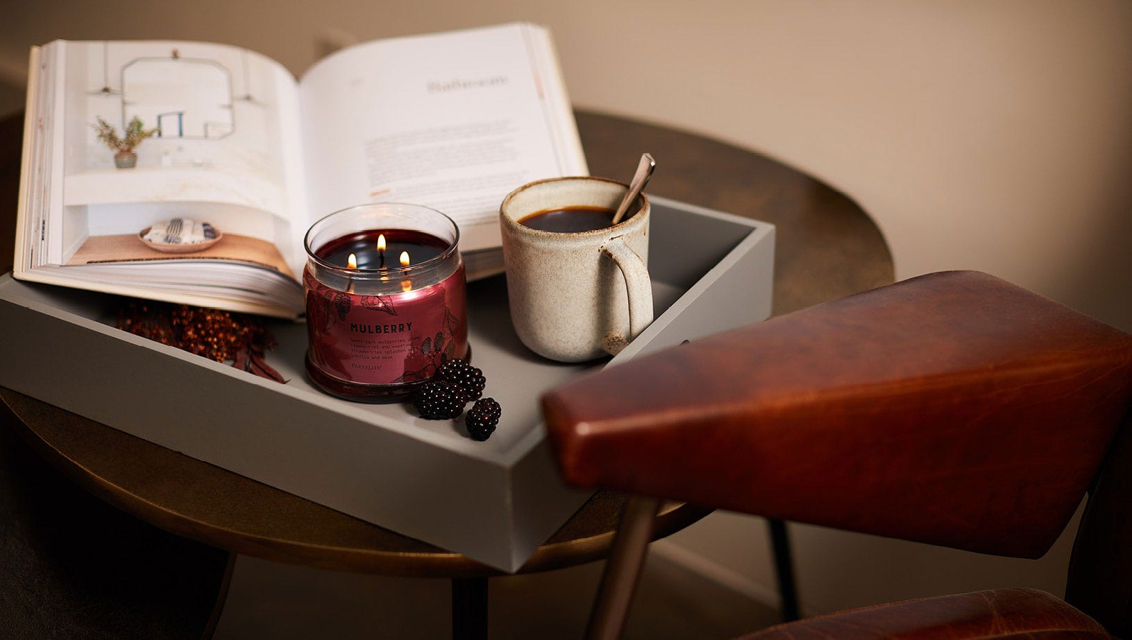 3-wick candle shown lit on a small table with a coffee cup and a book.