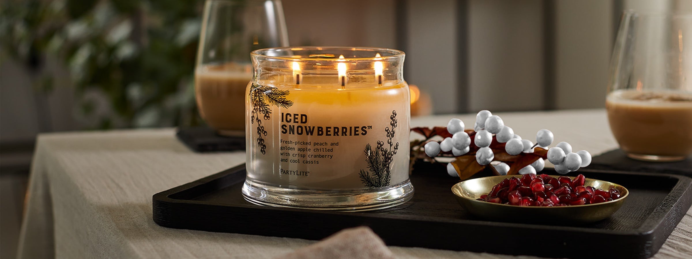 Lit off-white 3-wick candle on a tray next to red and white berries with two beverages in the background..