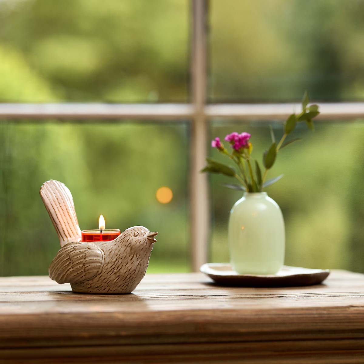 Songbird Tealight Holder on a wooden table with a flower vase with a window in the background