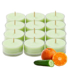 Twelve green tealight candles in glass holders with oranges and a cucumber on a white background.
