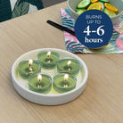 Round ceramic holder with five green tealight candles on a wooden table, with a bowl of fruit in the background, and a blue circle showing the burn times.