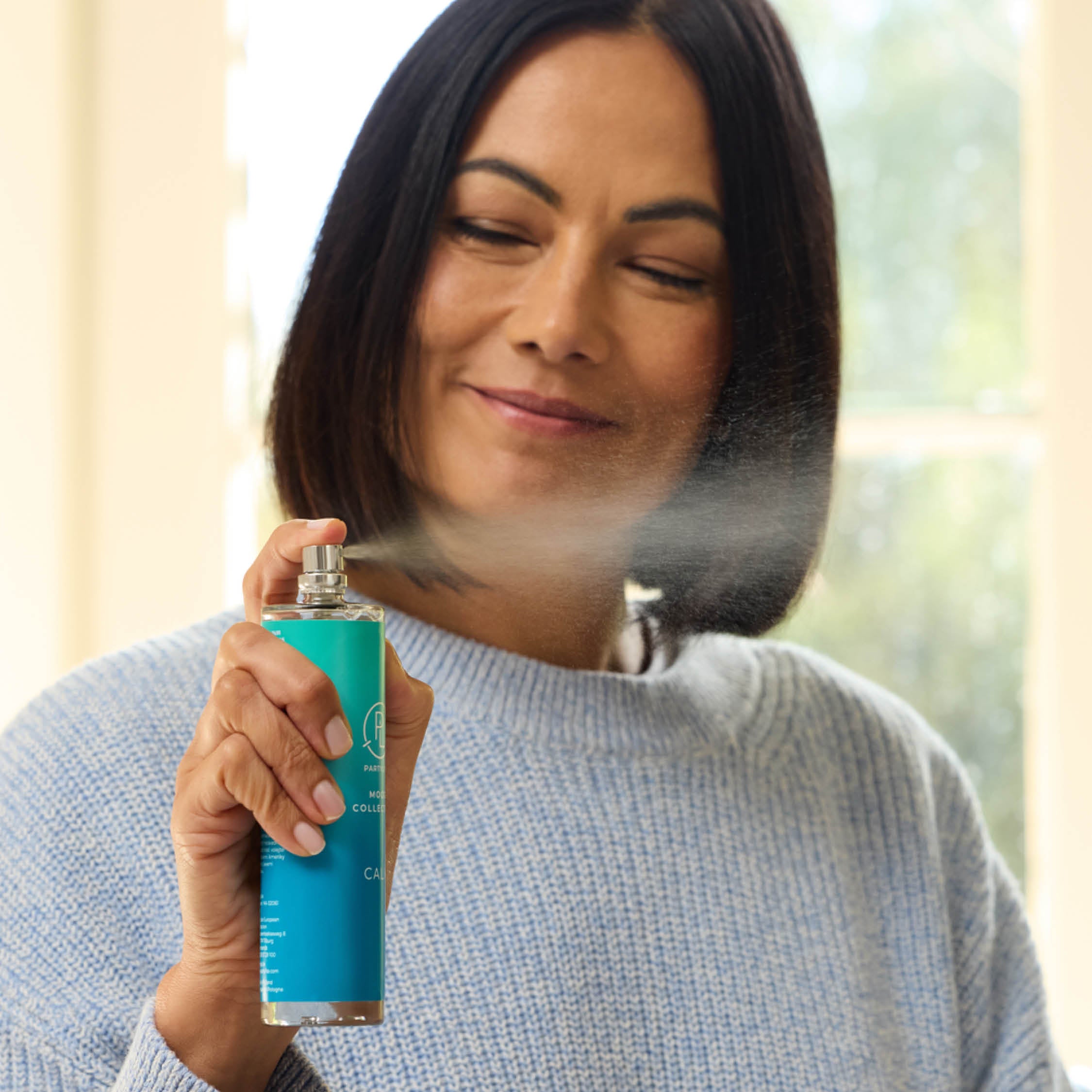 Woman holding a blue room spray bottle with mist coming out, wearing a light gray sweater.