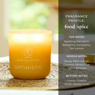 Yellow jar candle labelled 'Optimistic' with fragrance profile details on a blurred natural background.